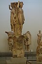 Photographie en couleurs de plusieurs statues de pierre, dont une dans les tons ocre figurant des femmes élancées ayant chacune le bras droit levé par dessus leur tête.