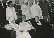 Sultan Muhammad V seated with Roosevelt and Churchill. Behind them, General Patton, Robert D. Murphy, Harry Hopkins, Hassan II, General Nogues, Muhammad al-Muqri, the Moroccan Chief of Protocol, Elliott Roosevelt, and John L. McCrea. Editing File-Franklin D. Roosevelt, Winston Churchill, Mohammed V and Hassan II at the Casablanca Conference.jpg