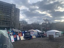 Several tents can be seen behind a long metal fence, covered in banners