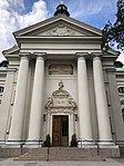Kyrkans huvudfasad med kopplade kolonner och fronton, samt entrén och inskriptionen på latin.