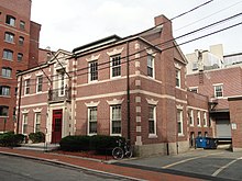 A small two story brick building with off white stone details at the corners, above the windows, and two string courses between the stories.