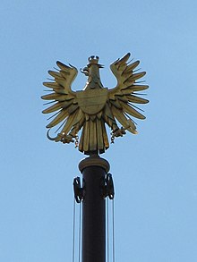 Künstlerisch stilisierte, gänzlich goldfarben ausgeführte Figur des Wappenadlers auf einem Flaggenmast vor dem Parlament