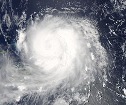 Satellite image of a slightly elongated tropical cyclone over blue waters; an eye, visible as a void at the center of the mass of white clouds, is visible. Small green islands dot the lower-left corner of the image.