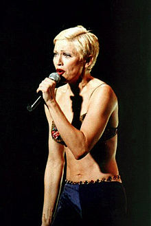 A woman with short blonde hair, wearing a green bra and purple pants, singing to a microphone, held in her left hand.