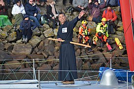 Debout sur le pont, en kimono noir et hakama noir (pantalon large et plissé), Kiraishi salue la foule. Un sabre de bois (bokutō, ou bokken, en japonais) est passé dans sa ceinture.