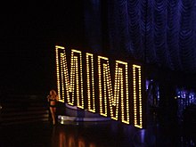 A still of Carey performing "Shake It Off" during The Adventures of Mimi Tour (2006). Carey is shown walking down the spiral staircase, onto the main stage, as the Broadway-styled 'MIMI' prop descend from the rafters. Mariah Carey in August 2006.jpg