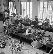 A view of the workroom at Henry Poole and Co., showing tailors at work on various types of jacket, including a naval officer's jacket, second from right on the rear row. The men are all sitting on the workbenches. (1944) Savile Row- Tailoring at Henry Poole and Co., London, England, UK, 1944 D21864.jpg