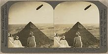 A pair of sepia images showing the Graf Zeppelin flying above the Great Pyramid. In the foreground, three people in Egyptian dress are sitting or kneeling with their backs to the camera, watching the scene. The airship's image is slightly further to the left on the right-hand image; when viewed through the correct apparatus this would give the illusion that it was further away.