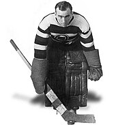 Black and white image of a hockey goaltender holding a stick, wearing pads and a horizontal=striped jersey