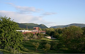 In der Ferne, umgeben von einem Feld und vor baumbewachsenen Hügeln, liegt ein Fabrikgebäude aus Backstein mit Schornstein