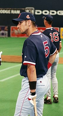 A man in white pants, a dark blue baseball cap, and a dark blue baseball jersey with "5" on the back leans on a baseball bat in his left hand.