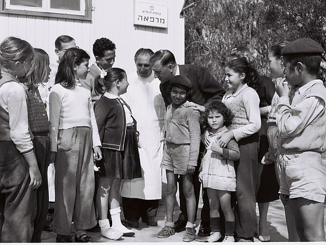 אורחים מהולנד פוגשים ילדים ישראליים בסניף קופת החולים של מעברת כפר סבא, בשנת 1958. הצילום בשחור ולבן, וכל האנשים בו לבושים בבגדים חגיגיים ומסודרים. לאחד המבוגרים יש חלוק של עובד רפואה.