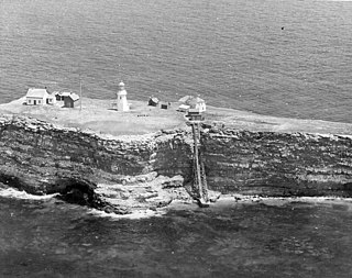 Photo du site, vue depuis un aéronef situé au sud. On y voit l'îlot : la falaise, le quai et l'escalier en bois, le phare avec la maison des gardiens et d'autres petits bâtiments.