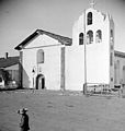 Mission Santa Inés in about 1912. The mission's original three-bell campanario, erected in 1817, collapsed in a storm in 1911 and was subsequently replaced by this concrete four-bell version, which also had openings on the side. This tower was replaced in 1948 to restore the original three-niched appearance. It has been compared by architectural historian Rexford Newcomb to the one that originally abutted the façade of Mission San Gabriel Arcángel.