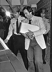 Tersenyum Bundy memegang setumpuk kertas dan masuk kendaraan. Ia dikawal oleh dua petugas polisi.