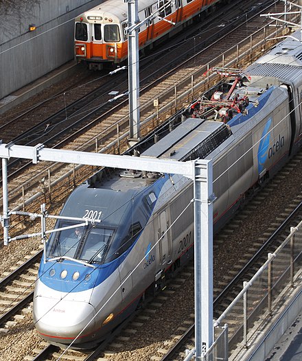 Overhead view of an Acela power car in Boston; an MBTA Orange Line subway train is also visible in the background.