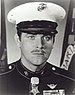 Head of a white man with a thin mustache wearing a white peaked cap with black visor and a dark military jacket with a medal hanging from a ribbon at the neck.