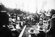 Men, women and children watch a stick game, where group of men sit in two lines, separated by 2x4s, some have sticks in hand. One man sits between the posts. Photo circa 1908. Colville stick game, Colville Reservation, ca. 1908.jpg