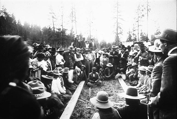 Men, women and children watch a stick game, where group of men sit in two lines, separated by 2x4s, some have sticks in hand. One man sits between the