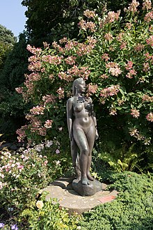 A photograph of a bronze sculpture of a nude woman holding a towel. The sculpture appears in a park or garden, in front of a lush, flowering bush with peach-colored flowers.