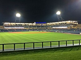 Una vista nocturna de la explanada de hormigón gris, los asientos azules y el campo de juego verde iluminado por dos postes de iluminación en la explanada después de un partido