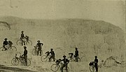 image en noir et blanc, 8 cyclistes chapeautés debout à côté de leur bicyclette devant des terrasses de travertin