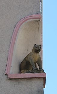 Bir binanın köşesinde bir niş içinde oturan bir kedinin yüksek heykeli.  Sağ ön dörtte üçünden görülüyor.