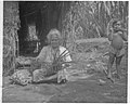 Photo en noir et blanc d'une femme assise âgée tressant des feuilles, à sa droite un enfant en short