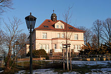 Villa Storchennest, a building with two floors and an attic under the red-roofed hipped roof, stands in a park-like setting.  The sand-colored facade is decorated with ornamental plastered surfaces in the Art Nouveau style.  The upper and lower floors are clearly separated from each other by a reddish cornice, and a bay window with a trapezoidal plan protrudes in the middle of the first floor.  A terrace in front of the ground floor is surrounded by red brick walls and has a railing made of white wooden slats.  In the middle of the roof, above the bay window, there is a larger dormer window with windows;  above it, on the roof ridge, there is a turret.  It's winter, the trees are bare and the wintry sun gives the facade a warm shade of yellow.