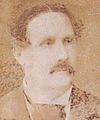 Verblasstes Sepiafoto, das den Kopf und die Schultern eines Mannes mit dunklem, welligem Haar, Schnurrbart und dunklem Mantel, weißem Hemd mit Kläppchenkragen und dunkler Krawatte zeigt