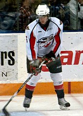 Un adolescent joueur de hockey sur glace manipulant une rondelle tout en restant immobile sur la glace.  Ses patins sont écartés de la largeur des épaules et ses yeux sont baissés.  Il porte un maillot blanc avec des garnitures rouges et noires, ainsi qu'un casque blanc à visière.