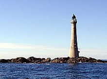 A tall lighthouse stands on low rocks under a blue sky.