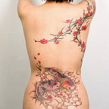 Femme nue de dos, avec des tatouages colorés de style japonais : branches de cerisier en fleur de l'épaule droite au milieu du dos, et deux carpes sur fond de gravier sur le haut du bassin.