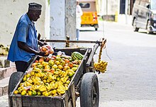 An African man sells produce out of a wheelbarrow containing mangos, oranges, watermelons, and the like.