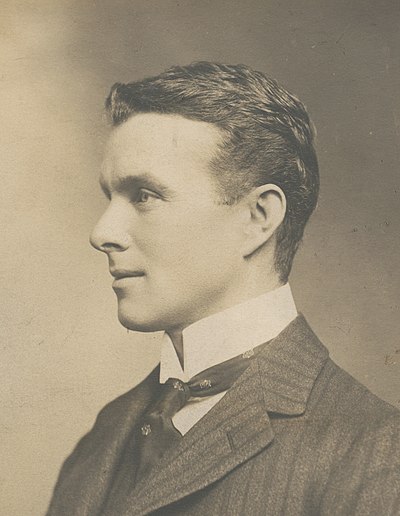 A man, fresh-faced with dark, brushed-back hair, seated among a group. He is wearing a naval officer