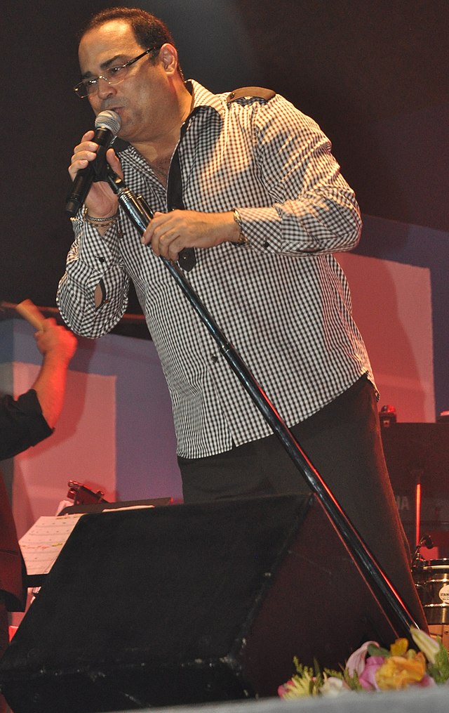 A man with glasses with a dark green patterned shirt is singing with a microphone on his right hand