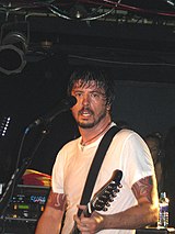 Un hombre con barba y camisa blanca toca la guitarra frente a un micrófono.