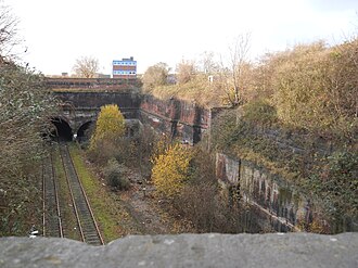 The three eastern portals of Liverpool Edge Hill tunnels, built into a hand dug deep cutting. The left tunnel with tracks is the short 1846 second Crown Street Tunnel, still used for shunting; next on the right partially hidden by undergrowth is the 2.03 km (1.26 mi) 1829 disused Wapping Tunnel, to the right again hidden by undergrowth, is the original short disused 1829 Crown Street Tunnel. Edge Hill cutting.jpg