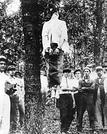 Leo Frank's corpse hanging from a tree after the lynching. His hands and feet bound. A crowd of spectators surrounds the tree.
