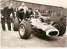 Graham Hill in BRM P261, testing at Silverstone in 1965. Chassis designer John Crosthwaite in duffel coat J Crosthwaite in duffel coat with Graham Hill in BRM.jpg