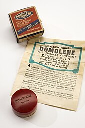 A jar of ointment, with a box and a poster. The box has the words "Domolene Brand Stops all skin troubles rashes and irritation The miracle ointment"