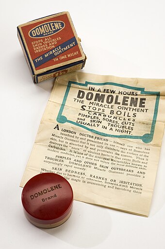 A jar of ointment, with a box and a poster. The box has the words 'Domolene Brand Stops all skin troubles rashes and irritation The miracle ointment'