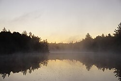 دریاچه Kinwamakwad (دریاچه طولانی) در صبح زود. خورشید در حال طلوع است اما هنوز قابل مشاهده نیست و یک لایه نازک از مه بخار بر روی سطح آب قرار دارد.