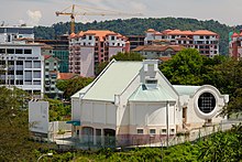 Fotografia de um grande edifício predominantemente branco.  É composto por uma parte em prisma triangular e outra em cilindro.  Está rodeado em primeiro plano por vegetação tropical, em segundo plano por edifícios e em segundo plano por montanhas.