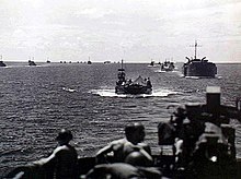 A group of World War II-era ships at sea photographed from another ship. Two men wearing helmets are in the foreground.