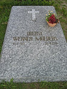 Colour photograph of a marbled-grey stone slab, lying flat on the ground, surrounded by grass and weeds, bearing the golden inscription "Oberst Werner Mölders 18.3.1913 22.11.1944" just below the centre of the slab. Above the inscription, there is a small Christian cross carved from similar, but slightly lighter stone, and a terracotta bowl with flowers near the right-hand edge of the slab.