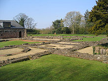 Low stone walls form squares and other shapes and are surrounded by grassed areas. In the background to the left is the rear of the undercroft, and trees are in the background.