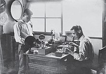 Dos hombres trabajando en una mesa con varios dispositivos electrónicos.  Uno está parado en el lado izquierdo de la mesa.  El otro, sentado en el lado derecho de la mesa, usa audífonos y prueba una radio de cristal.