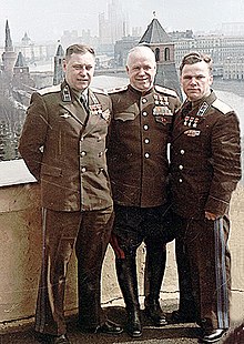 Photo en couleur d'un groupe de trois hommes en uniforme et portant médaille, sur un balcon au-dessus de la place rouge.