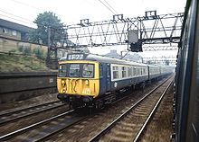 The front of a British Railways Class 312 stock train in British Rail corporate 1970s Blue/Grey livery passes through East London on a working to London's Liverpool Street station, as seen from the open window of another train 312-seen-out-train-window.jpg